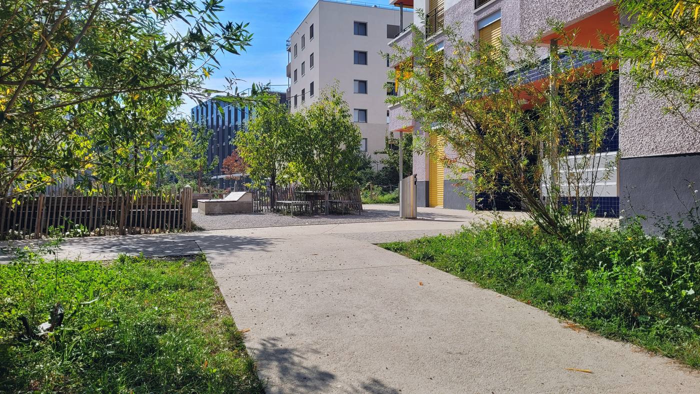 Cheminement en béton menant à une placette en stabilisé avec tables
